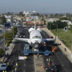 A space shuttle is transported through a city on a multi-wheeled vehicle, surrounded by people and vehicles, with buildings and trees in the background.