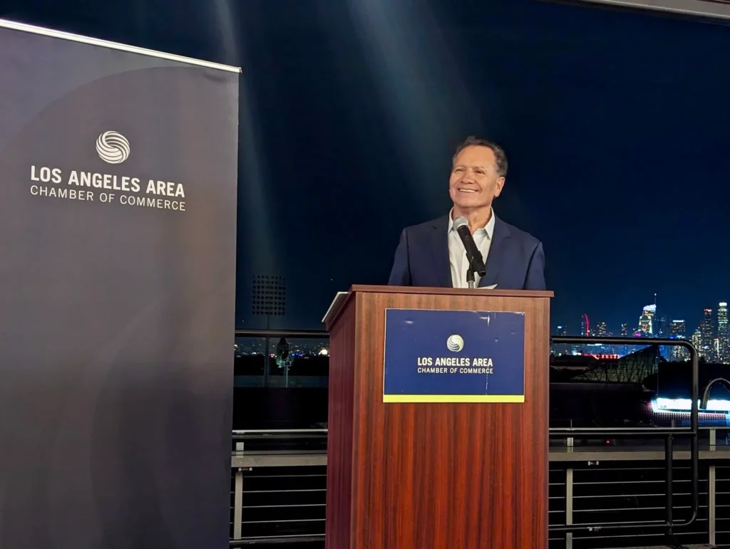 A smiling man in a navy blue suit stands at a podium with a microphone, delivering a speech at a Los Angeles Area Chamber of Commerce event. A banner with the organization's logo is displayed to the left, and the illuminated Los Angeles skyline serves as the backdrop.