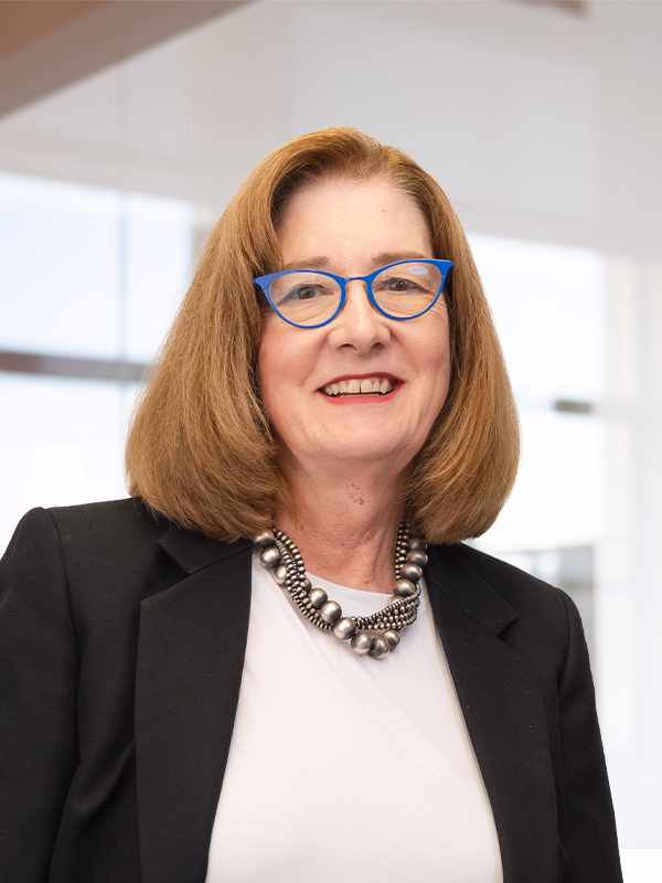 Professional headshot of a smiling woman with red hair, blue glasses, black blazer, and statement necklace in modern office setting