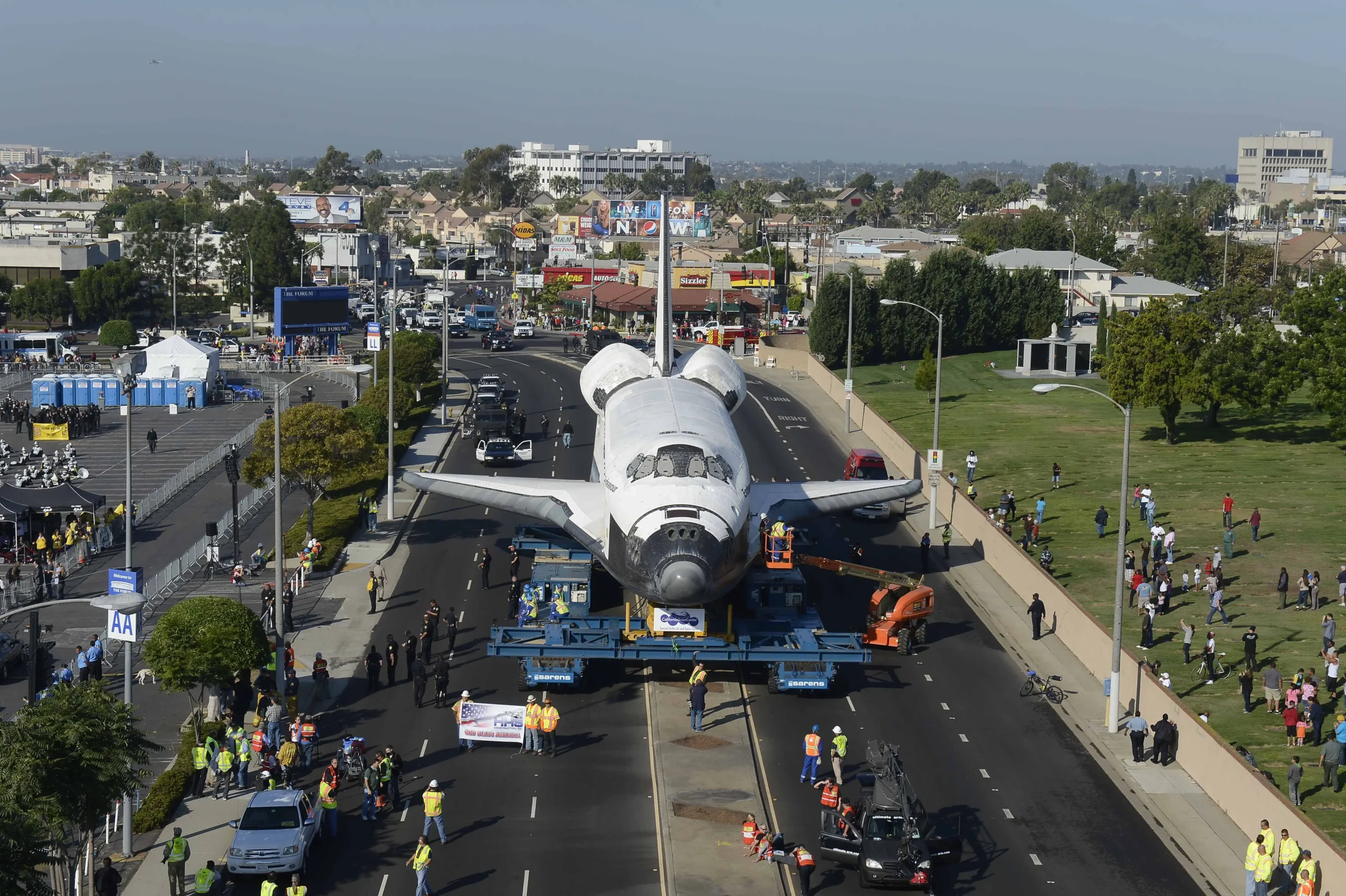 A space shuttle is transported through a city on a multi-wheeled vehicle, surrounded by people and vehicles, with buildings and trees in the background.