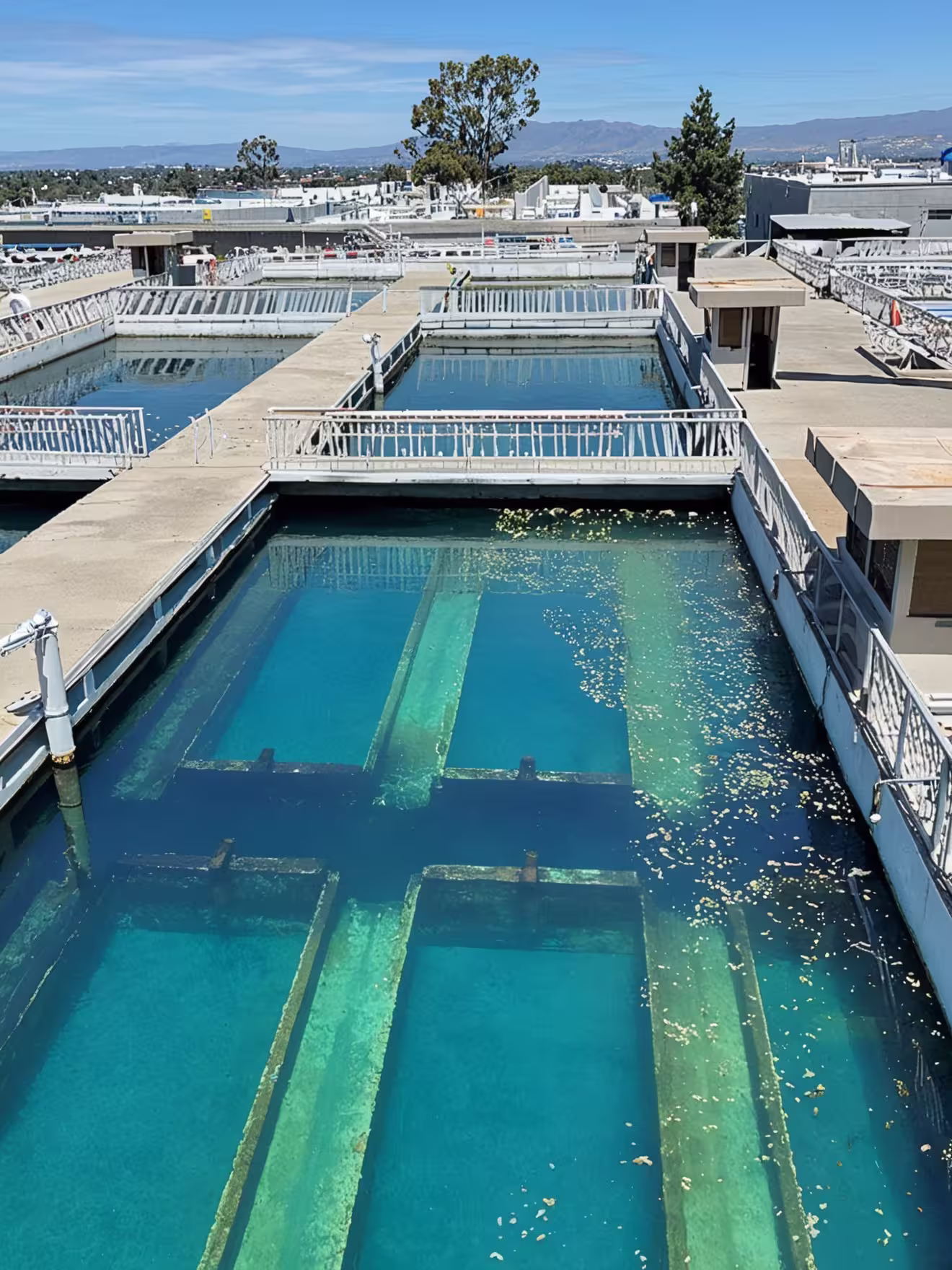 Large industrial water treatment tanks with clear blue water, concrete walkways, and metal railings under a sunny sky.