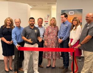Group of diverse professionals standing behind a red ribbon at a ribbon-cutting ceremony in an office setting, with a banner in the background.